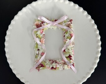 May include: A small, rectangular, decorative frame made of textured yarn in shades of pink, green, and white, with a pink ribbon tied in a bow. The frame sits on a white, scalloped-edge plate against a black background.