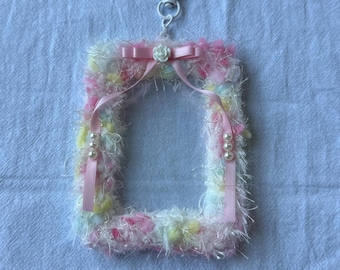 May include: A rectangular, fluffy keychain frame in pastel colors of pink, yellow, and white. The frame is adorned with a pink ribbon, a small white rose, and faux pearls. A white clasp and a pearl necklace are attached.