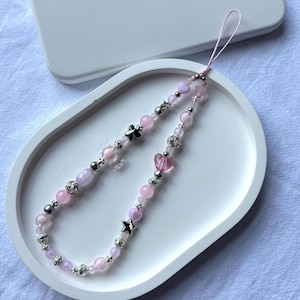 Puede incluir: Un charm de teléfono con cuentas que presenta cuentas rosas y lavanda, detalles plateados y dijes de corazón y estrella. El charm se muestra en una bandeja ovalada blanca, con una funda de teléfono blanca en el fondo.