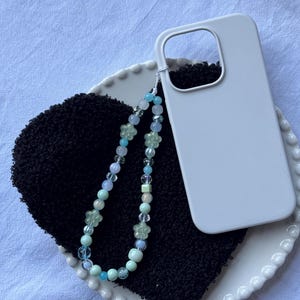 Puede incluir: Una funda de teléfono blanca descansa sobre una superficie negra texturizada, junto a una correa de teléfono con cuentas. La correa presenta cuentas transparentes, azules y verdes, incluyendo detalles en forma de flor. Los artículos están sobre un plato blanco festoneado.