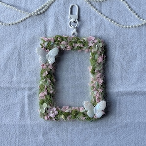 Op de afbeelding: Een rechthoekige badgehouder met een bloemig thema, een witte sluiting en een parelketting. Het frame is versierd met groene en roze bloemaccenten en twee witte vlinderversieringen. De badgehouder is ontworpen om een ID of kaart te bevatten.