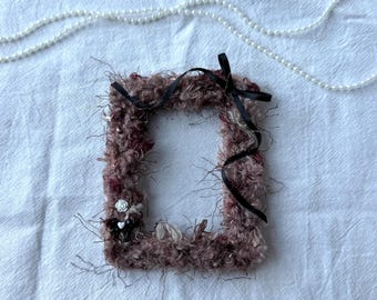 May include: A rectangular, fuzzy, mauve-colored picture frame with a black ribbon and small white flower accents. A white pearl necklace is in the background. The frame has a textured, handmade appearance.