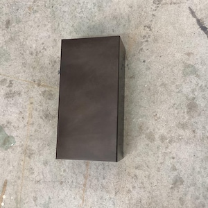 May include: A rectangular, dark brown box with a matte finish. The box is positioned upright on a concrete surface. The box appears to be made of metal or a similar material. The overall aesthetic is minimalist and industrial.