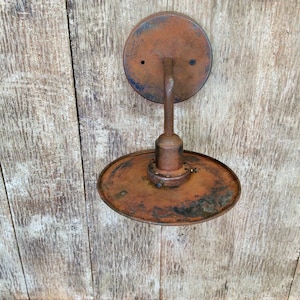 May include: Vintage wall-mounted light fixture with a circular backplate and a downward-facing, oval shade. The fixture is made of rusted metal, showing signs of age and wear. The overall design is industrial.