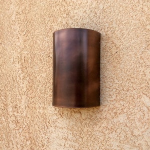 May include: A copper-colored wall sconce with a rounded rectangular shape. The sconce is mounted on a textured wall.