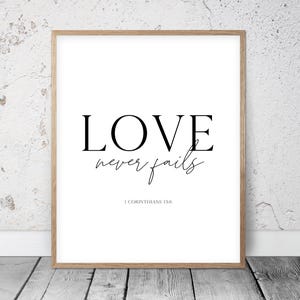 Op de afbeelding: Een ingelijste print met de woorden "LOVE never fails" in zwarte letters op een witte achtergrond. De tekst "1 CORINTHIANS 13:8" staat eronder. De lijst is lichtbruin.