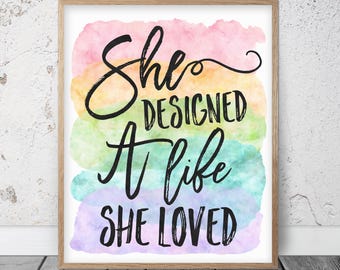 以下が含まれることがあります： 「She Designed A Life She Loved」という引用句が黒い筆記体で書かれた額入りのアートプリント。背景はピンク、黄色、緑、青、紫の色合いの水彩画。フレームは明るい茶色です。