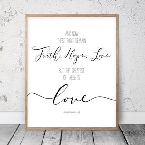 Pode incluir: Uma impressão emoldurada com as palavras "Fé, Esperança, Amor" em escrita elegante. O texto "E agora, permanecem estas três" e "Mas o maior destes é o amor" também são exibidos. O versículo bíblico 1 Coríntios 13:13 está na parte inferior.