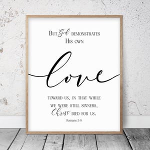 Puede incluir: Una impresión enmarcada con fondo blanco y texto negro. El texto dice: "Pero Dios demuestra su amor por nosotros, en que siendo aún pecadores, Cristo murió por nosotros". El marco es de color marrón claro.