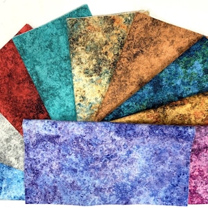 May include: A collection of textured fabric swatches in various colors, including red, teal, silver, blue, and gold. The fabric has a mottled appearance, with darker speckles on a lighter background. The swatches are arranged in a fan shape.