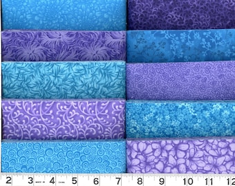 May include: A collection of ten folded fabric strips in shades of blue and purple, featuring various floral and swirling patterns. A white measuring tape is visible along the left and bottom edges, showing measurements in inches up to 21 inches.