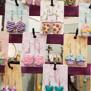 May include: A display of handmade polymer clay earrings featuring various food and Easter-themed designs. The earrings are displayed on white cards with black hooks. Some of the designs include a pizza slice, a cupcake, a Peep, and a breakfast sandwich.