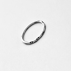 May include: A silver ring with a repeating pattern of small, dark ovals. The ring is thin and has a smooth, polished surface. The design is simple and elegant, suitable for everyday wear. The ring is shown against a white background.