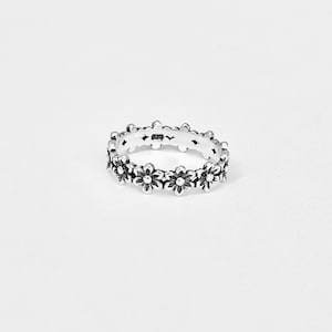 May include: A silver ring with a daisy flower pattern. The ring has a repeating design of small flowers around the band. The ring is made of a shiny metal and is set against a white background.