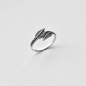 May include: A silver-colored ring featuring a leaf design. Two detailed leaves are positioned on the top, with a band adorned with small beaded accents. The ring is set against a white background.