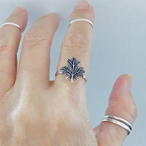 May include: A silver ring featuring a detailed maple leaf design. The ring is worn on a finger, showcasing the intricate leaf pattern. Additional silver rings are visible on other fingers, set against a light background.