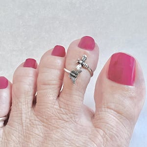 Op de afbeelding: Een zilveren teenring met bijen- en vlindermotieven. De ring wordt gedragen op een voet met magenta nagellak. Het ontwerp van de ring toont een bij aan de bovenkant en een vlinder aan de zijkant. De ring is verstelbaar.