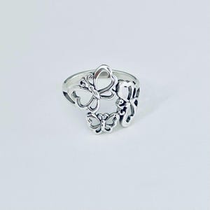 May include: A silver-toned ring featuring an openwork design of three connected butterflies. The metal has a polished finish with darkened recessed areas to define the wing patterns and body shapes of the insects. The band is smooth and circular.
