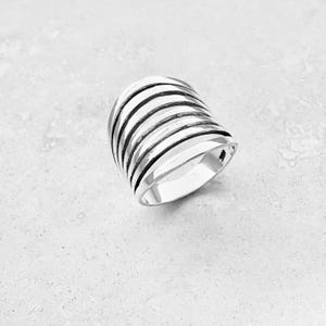 May include: A silver ring with a wide band made of multiple stacked, curved, and slightly textured bands.