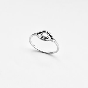 May include: A silver-colored ring featuring an eye design. The ring has a thin band and a stylized eye with a circular center. The eye is outlined with a darker shade, creating a contrast against the lighter band. This ring is a piece of jewelry.