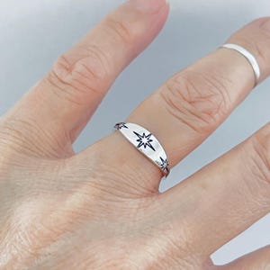 May include: A silver ring with a starburst design. The ring features a central starburst and smaller stars along the edges. The ring is worn on a finger.