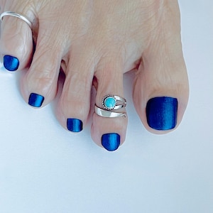 May include: A silver-toned toe ring featuring a small, round, bright turquoise stone set within a twisted rope-style border. The band is open and adjustable, wrapping around the second toe, while a plain silver band is worn on the big toe.