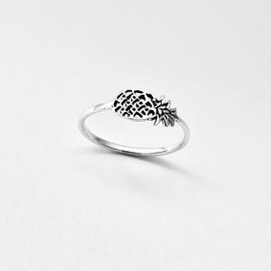 Könnte beinhalten: Ein silberner Ring mit einem Ananas-Motiv. Die Ananas hat eine strukturierte Oberfläche und eine detaillierte Krone. Der Ring wird vor einem weißen Hintergrund präsentiert, um das Design des Schmuckstücks hervorzuheben.