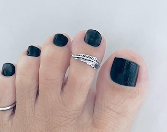 May include: A silver toe ring with a feather design. The ring is on a person's toe with black nail polish.