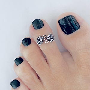 Puede incluir: Un anillo para el dedo del pie de metal plateado con un delicado diseño de hojas caladas. El anillo se lleva en el segundo dedo del pie, situado junto al dedo gordo con esmalte de uñas negro oscuro. La banda es fina y rodea el dedo.