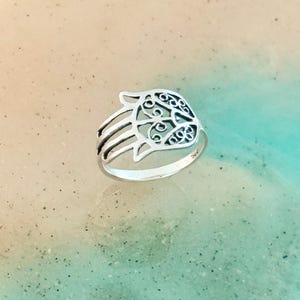 May include: A silver ring with a Hamsa hand motif. The openwork hand design has intricate patterns within the palm and fingers. The ring is set against a backdrop of neutral and turquoise tones, creating a visually appealing contrast.