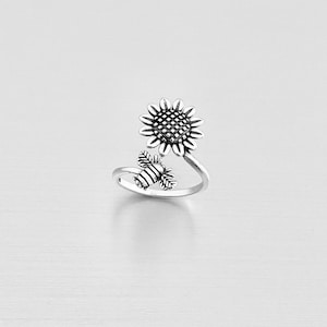 May include: A silver-toned ring featuring a sunflower and a bee design. The sunflower is at the top, with detailed petals and a textured center. The bee is positioned below, with striped detailing. The ring's band curves around to create the design.