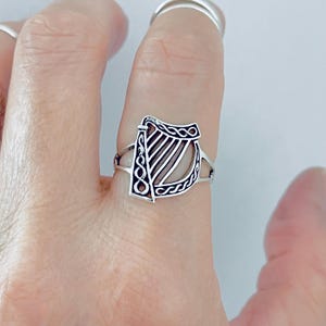 May include: A silver-toned ring featuring a harp-shaped centrepiece with intricate cut-out details and a braided pattern along the frame. The band is thin and split, connecting to the sides of the harp design, creating a balanced and open appearance.