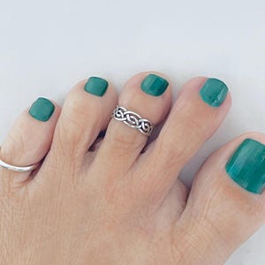 Puede incluir: Anillo de dedo del pie de plata con un diseño trenzado. El anillo está en un pie con esmalte de uñas verde.