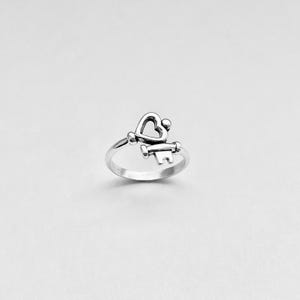 Puede incluir: Un anillo de plata con un diseño de llave en forma de corazón. La llave está abierta y el corazón es hueco.