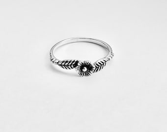 Könnte beinhalten: Ein silberner Ring mit floralem Design. Der Ring hat eine zentrale Blume mit detaillierten Blütenblättern, flankiert von Blättern auf beiden Seiten. Das Band ist glatt und kreisförmig, wodurch ein zartes, von der Natur inspiriertes Schmuckstück entsteht.