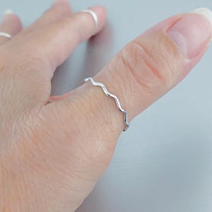 May include: A thin, silver-colored ring with a continuous wavy or serpentine design worn on a finger. The band features a smooth, reflective metallic finish and a consistent undulating pattern that wraps around the circumference of the finger.