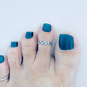 May include: A close-up of a foot with teal nail polish and a silver toe ring. The toe ring features a wave design. The foot is against a white background.