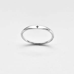 May include: A thin, polished silver-tone ring featuring a small, centered black cross symbol engraved on the top surface. The band has a smooth, rounded profile and a reflective metallic finish, creating a simple and clean appearance.