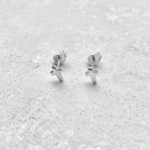 May include: A pair of small, polished silver stud earrings shaped like crosses. Each earring features a simple cross design with a post back, showcasing a bright, reflective metallic finish that catches the light on its smooth, clean surfaces.