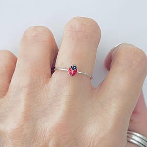 May include: A thin silver ring featuring a small, enamel ladybug charm. The ladybug is painted red with black spots and a black head. The band is a simple, polished silver wire that encircles the finger, providing a delicate and understated appearance.