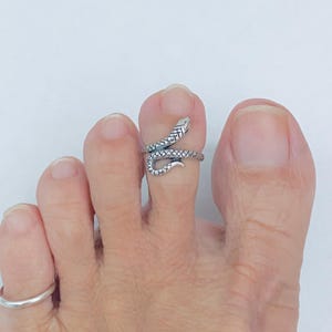 Könnte beinhalten: Ein silberfarbener Zehenring in Form einer Schlange, die sich um eine Zehe windet. Der Schlangenkopf befindet sich oben an der Zehe, und der Ring wird am zweiten Zeh getragen. Der Ring hat ein strukturiertes, detailliertes Design.