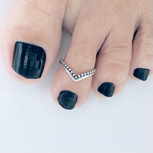 May include: A silver toe ring with a V-shaped design and beaded edges. The ring is worn on a toe with black nail polish.