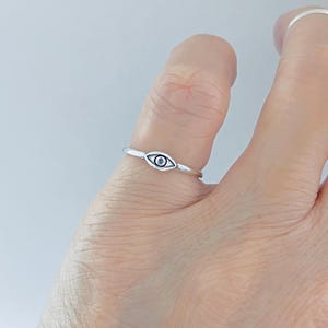 May include: A thin silver-toned ring featuring a small, almond-shaped eye motif at the center. The eye design includes a dark, circular pupil and a defined outline, set on a simple, slender band that encircles the finger.