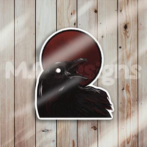 Può includere: Un corvo nero con un occhio bianco è in silhouette su uno sfondo rosso a forma di sole. Il becco del corvo è aperto e sembra star chiamando.