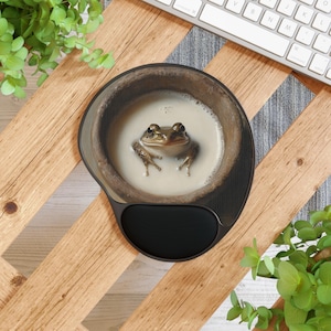 May include: A brown frog sits in a ceramic bowl filled with a creamy white liquid. The bowl is on a black and brown mouse pad.