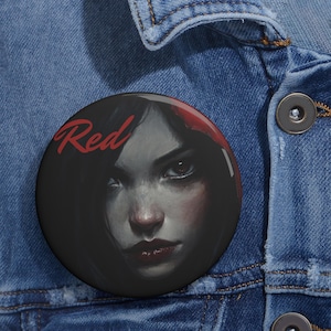 May include: A round button with a black and red design. The button features a woman's face with dark hair and red lips. The word "Red" is written in red letters on the button.