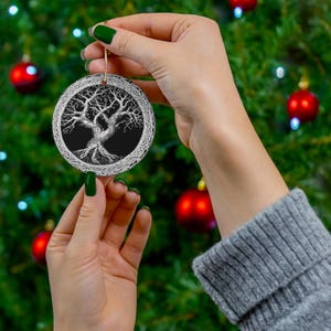 May include: A round Christmas ornament with a black background and a white detailed drawing of a tree with roots and branches. The ornament is being held by a hand.