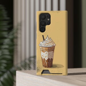 以下が含まれることがあります： ホイップクリームを添えたコーヒー飲料の漫画イラストが描かれた黄色の携帯電話ケース。ケースにはカメラモジュールと黒いストローが付いています。背景は薄茶色です。