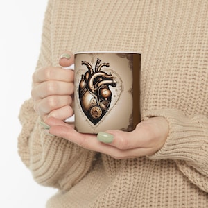 May include: A white ceramic mug with a brown and gold design of a steampunk heart. The heart is made of gears and cogs and has a clock face in the center.