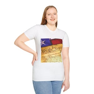 May include: White t-shirt with a graphic depicting a flag design, including a blue star, red and blue stripes, and the text "We the People" over a parchment background. The shirt has a crew neck.